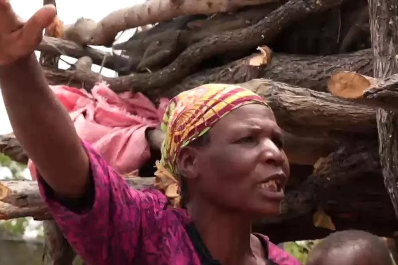 A woman speaking to camera beside stacked firewood during a community interview