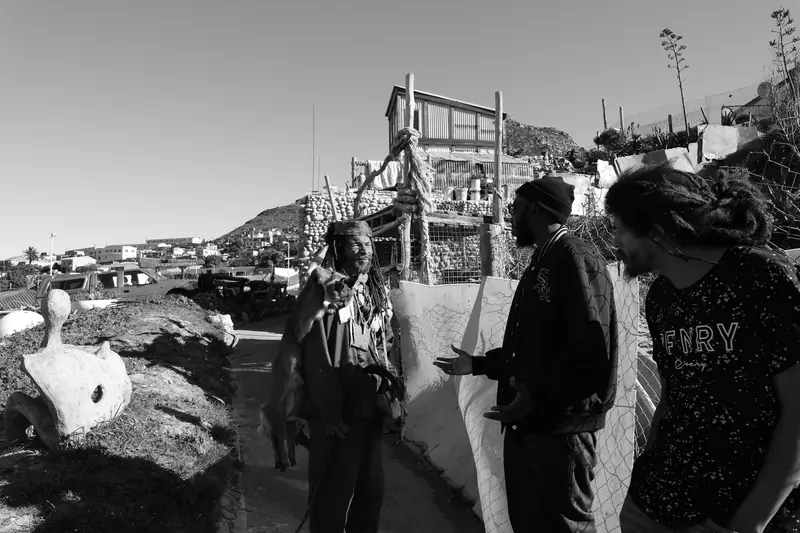 Three people in conversation on a hillside among informal structures, photographed in black and white