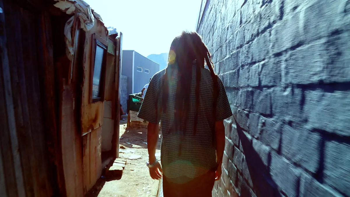 A figure walks a narrow lane between corrugated iron and brick walls in Hangberg
